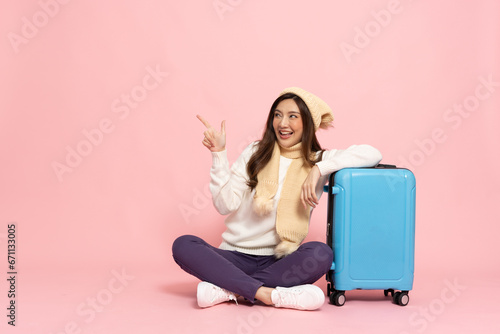 Happy Asian woman traveler sitting with luggage and pointing to copy space isolated on pink background, Tourist girl having cheerful holiday trip concept, Full body composition photo