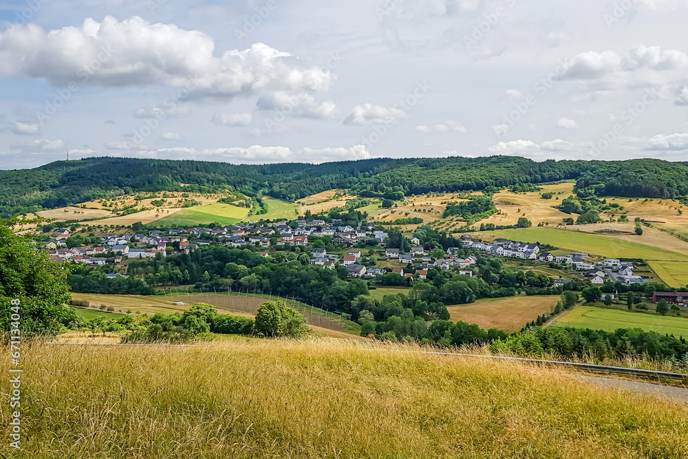 Panoramic landscape of German countryside with small village and farmland, green leafy trees covering a hill in background, sunny day in Eiffel Kreis, Bitburg-Prüm district in Rijnland-Palts, Germany