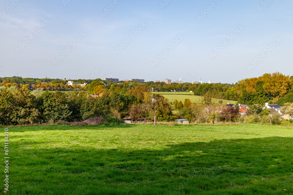 Plain of an agricultural plots with autumn trees with yellowish green foliage, city buildings against blue sky in background, sunny day in Sweikhuizen, South Limburg, Netherlands