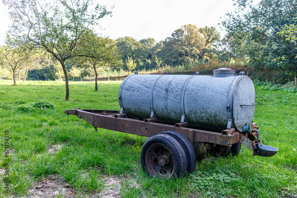 Old mobile water tank or waterer for livestock on an agricultural plot on Dutch farm, trees with green foliage in background, sunny day in Sweikhuizen, South Limburg, Netherlands Old mobile water tank or waterer for livestock on an agricultural plot on Dutch farm, trees with green foliage in background, sunny day in Sweikhuizen, South Limburg, Netherlands