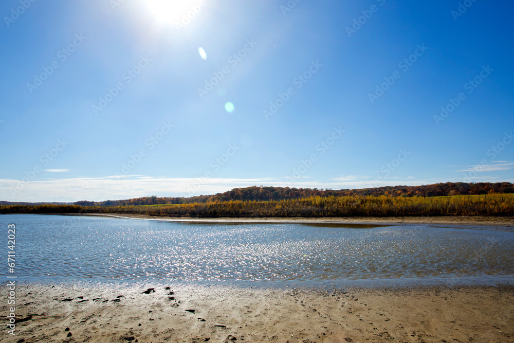 Autumn landscape of a lake and fall foilage in the background with sun and sun flares in the sky. 