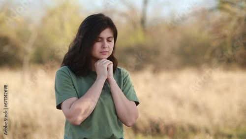 young woman folded hands and lowered her head in prayer to God standing on background of autumn nature, religion and faith concept
