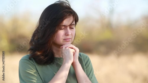 young woman folded hands and lowered her head in prayer to God standing on background of autumn nature, religion and faith concept
