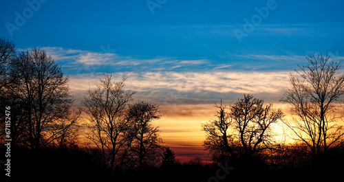 A serene sunrise over a field  with silhouetted trees in the foreground and a sky painted with hues of orange and blue. Silhouettes of trees against the setting sun. Evening landscape.