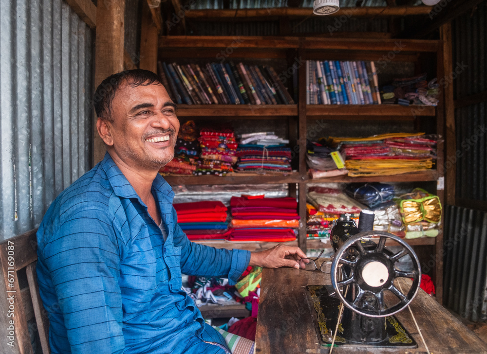 Smiling portrait of a south asian rural tailor sitting in front of his old fashioned sewing machine inside his small clothing store 