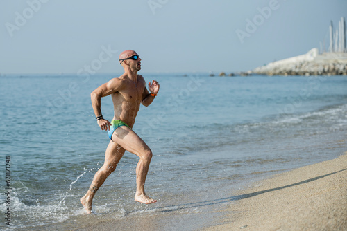Side view of a sexy muscular man in colorful speedo running out of the sea. Professional male athlete getting out of the water wearing swim glasses at the beach.