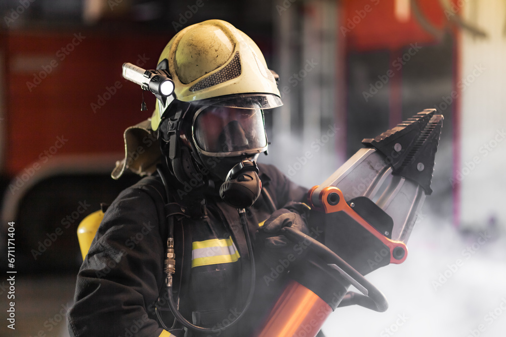 Fireman portrait wearing full equipment and oxygen mask. Firefighter with power hydraulic cutting tool. Smoke and fire trucks in the background. Fireman portrait wearing full equipment and oxygen mask. Firefighter with power hydraulic cutting tool. Smoke and fire trucks in the background.