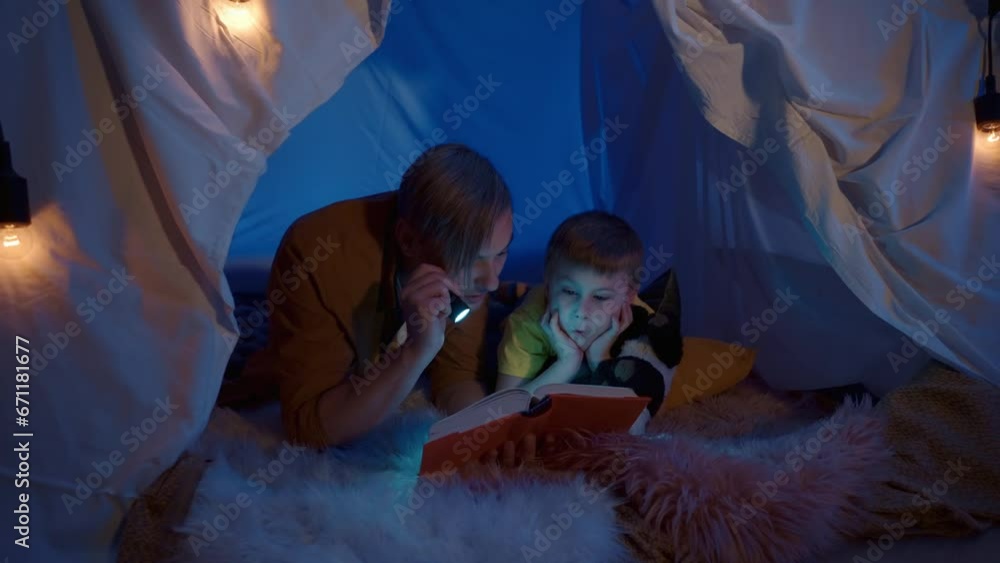 Dad and son reading a book in a tent in a dark room. A man and a boy lie on their bellies on a soft fur plaid, the man holds a flashlight pointing it at the book, the son nearby listening and yawning. Dad and son reading a book in a tent in a dark room. A man and a boy lie on their bellies on a soft fur plaid, the man holds a flashlight pointing it at the book, the son nearby listening and yawning.