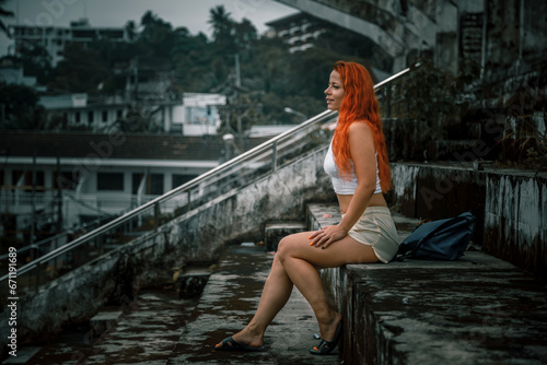 A beautiful girl sits in the stands of an abandoned stadium. It's a nasty day. Dilapidated stands. Red hair. Fan girl.
