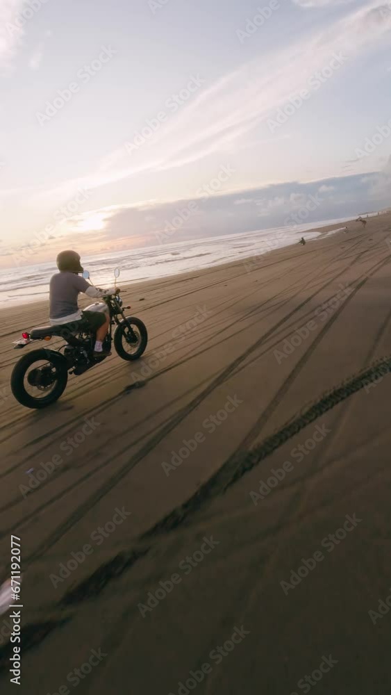 Vertical video. FPV sport drone following cafe racer motorcyclist biker riding tropical sandy beach. Guy in helmet enjoying driving custom motorcycle ocean shore sunset extreme professional bike sport Vertical video. FPV sport drone following cafe racer motorcyclist biker riding tropical sandy beach. Guy in helmet enjoying driving custom motorcycle ocean shore sunset extreme professional bike sport