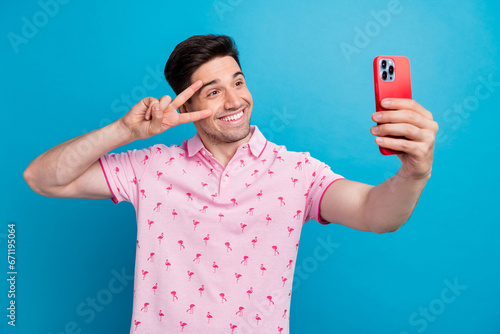Photo of positive young man hold smart phone make selfie show v-sign near eye isolated on blue color background