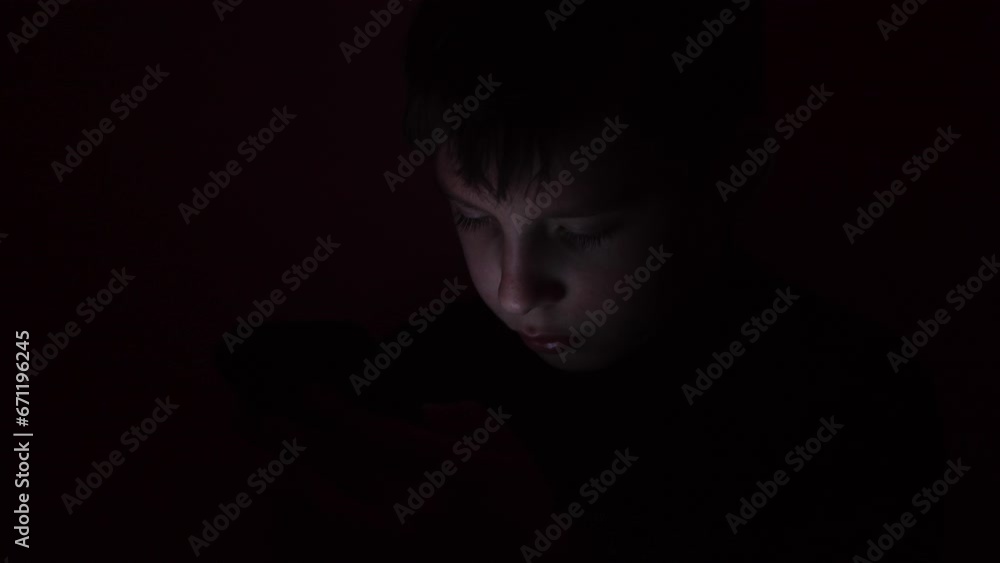 A little boy talks on a mobile phone under a blanket. Curious boy sitting in bed and playing games on a smartphone in a dark room. Image of a cute boy in pajamas sitting in front of a mobile phone. A little boy talks on a mobile phone under a blanket. Curious boy sitting in bed and playing games on a smartphone in a dark room. Image of a cute boy in pajamas sitting in front of a mobile phone.