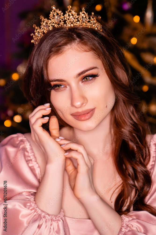 Close-up portrait of a young woman in a pink dress with a crown on her head posing against the background of a Christmas tree.