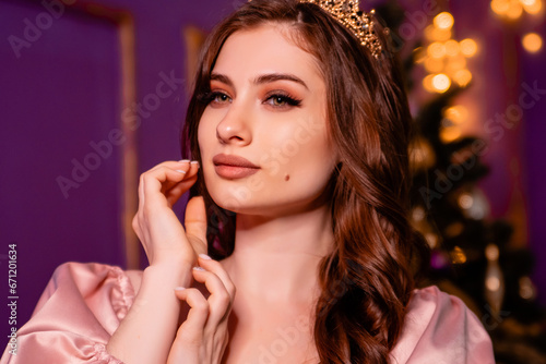 Close-up portrait of a young woman in a pink dress with a crown on her head posing against the background of a Christmas tree.