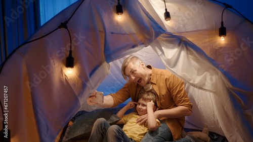 A father and son sit on fur skins in a tent in a dark living room and use a smartphone. Man and boy take selfies together, making funny faces, smiling, close up.