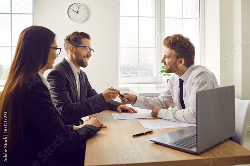 Real estate agent gives a contract and a pen to the clients. Young couple signing a purchase agreement at the realtor's office. Happy man and woman sitting at a desk with a friendly estate agent