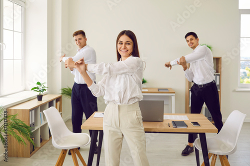 Fototapeta Naklejka Na Ścianę i Meble -  Happy corporate business team having fitness workout in office. Group of three people doing sports exercises. Smiling young woman and two men standing in office and doing shoulder stretching movements