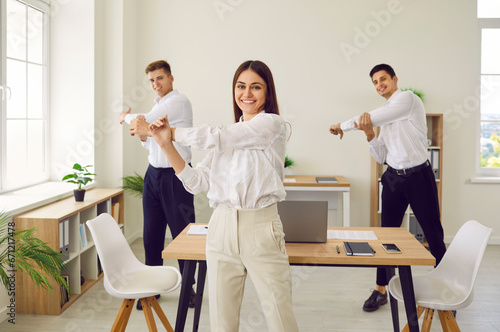 Happy corporate business team having fitness workout in office. Group of three people doing sports exercises. Smiling young woman and two men standing in office and doing shoulder stretching movements photo