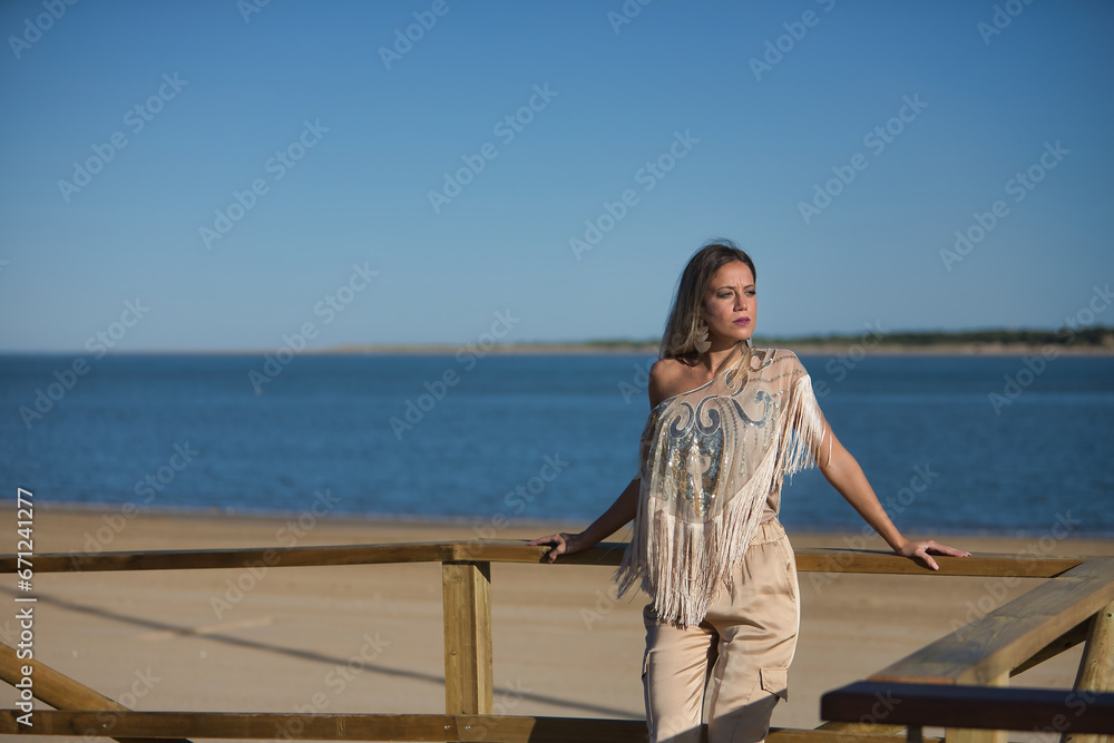Young and beautiful woman with sequined and fringed shirt, leaning on a wooden railing, looking at infinity by the sea. Concept beauty, fashion, trend, empowerment.