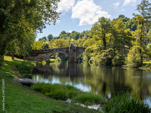 Vista del puente romano sobre el río tranquilo en Allaritz, rodeado de árboles verdes cielo azul y nubes blancas, en Orense, viajando por Galicia España en verano de 2021 photo