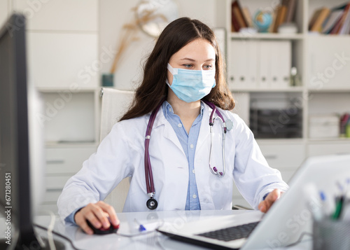 Woman doctor in protective mask sitting at workplace with computer in her office