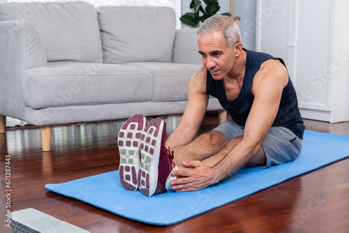 Active and fit senior man warmup and stretching before home exercising routine at living room while watching online fitness video. Healthy lifestyle concept after retirement for pensioner. Clout
