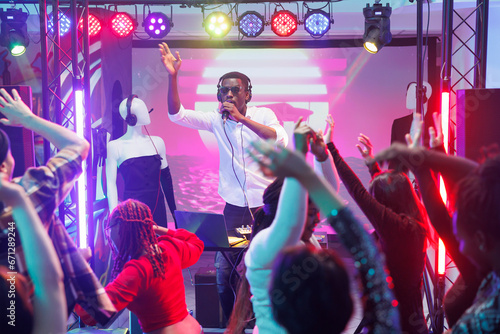 African american dj performing on stage and speaking to dancing crowd in microphone. People relaxing and moving on dancefloor at electronic music discotheque party in night club