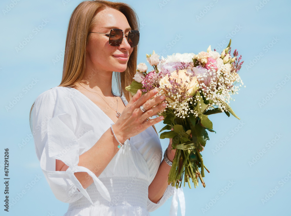 Joyful Young Woman Holding a Bouquet of Flowers on a Beach with Blue Sky Background