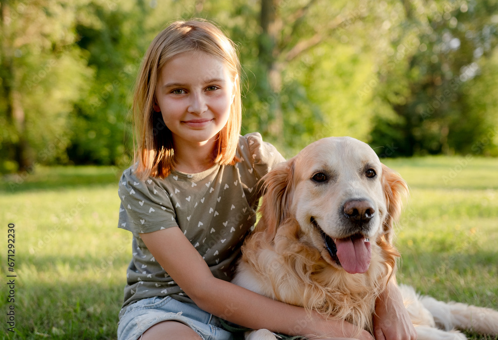 Cute preteen child girl hugging golden retriever dog at nature and sitting at grass. Pretty kid with purebred doggy pet labrador at park