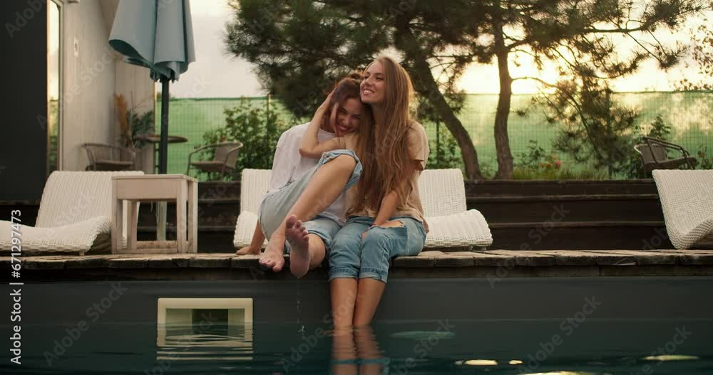 Two girlfriends are sitting near the sunbeds by the pool, hugging and looking into the distance. Rest in the country house Two girlfriends are sitting near the sunbeds by the pool, hugging and looking into the distance. Rest in the country house