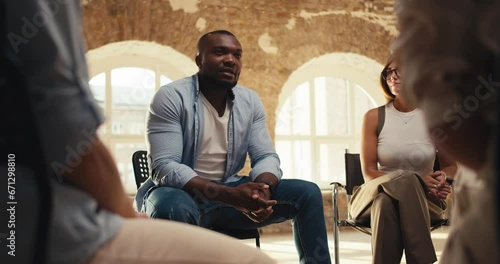 A Black man in a blue shirt expresses his thoughts during group therapy in a brick room with yellow lighting. A girl in a white T-shirt and glasses listens attentively to a man