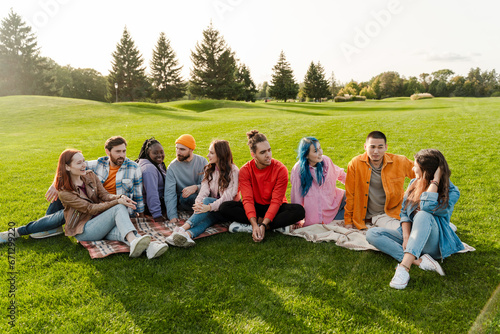 Group of smiling attractive multiracial friends wearing colorful clothing meeting, talking, relaxing, chilling, sitting on green grass in park. Diversity, friendship, travel concept  © Maria Vitkovska