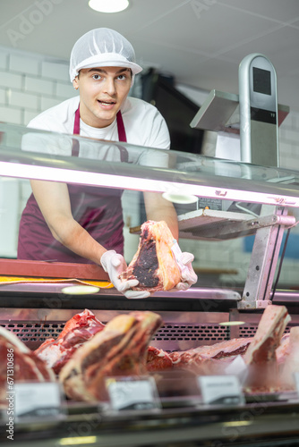 Sales guy in meat department of supermarket makes out showcase and puts pieces of meat on bones on shelf. Red meat, poultry, meat products and semi-finished products