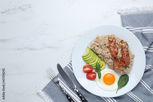 Delicious boiled oatmeal with fried egg  bacon  avocado and tomato served on white marble table  flat lay. Space for text