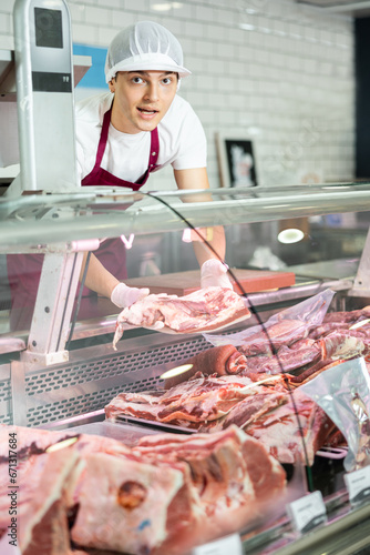 Courteous young shop assistant standing at showcase with big piece of row meat in hands