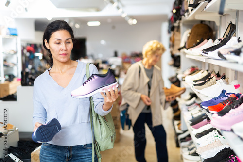 Fototapeta Naklejka Na Ścianę i Meble -  Asian woman selecting new sport shoes while standing in salesroom of shoeshop. Elderly European woman shopping in background.
