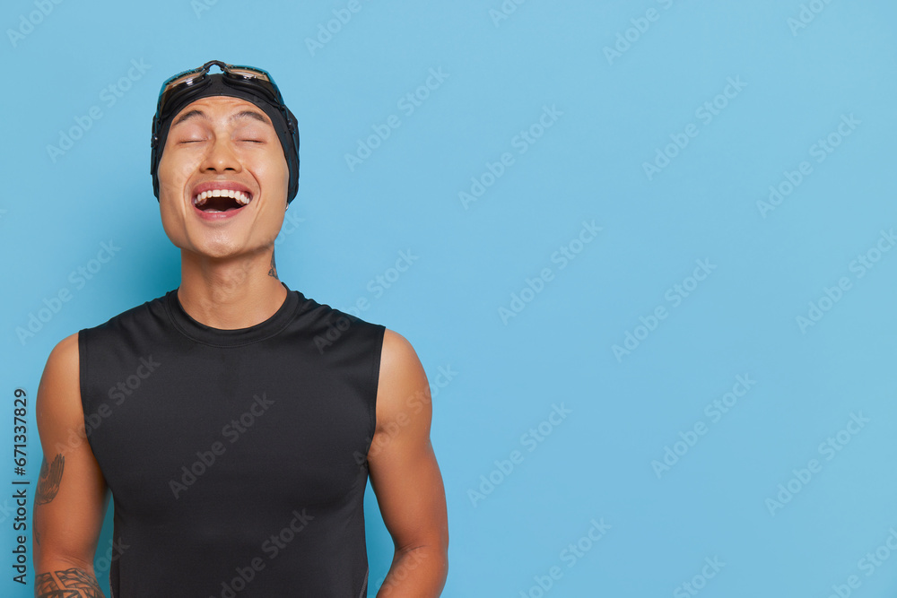 Young asian boy wearing black sleeveless top and swimming cap with goggles laughing over blue backdrop, sport life concept, copy space