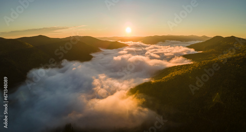 Aerial view of bright foggy morning over dark mountain forest trees at autumn sunrise. Beautiful scenery of wild woodland at dawn