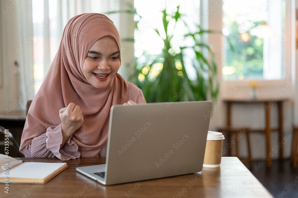 A cheerful Asian-Muslim woman is looking at her laptop screen with a happy face, good news. A cheerful Asian-Muslim woman is looking at her laptop screen with a happy face, good news.