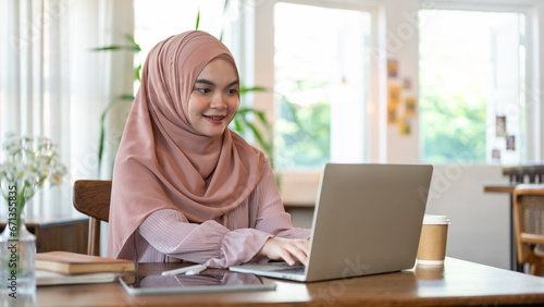 A happy Asian-Muslim woman is working remotely at a coffee shop, managing her work on her laptop.