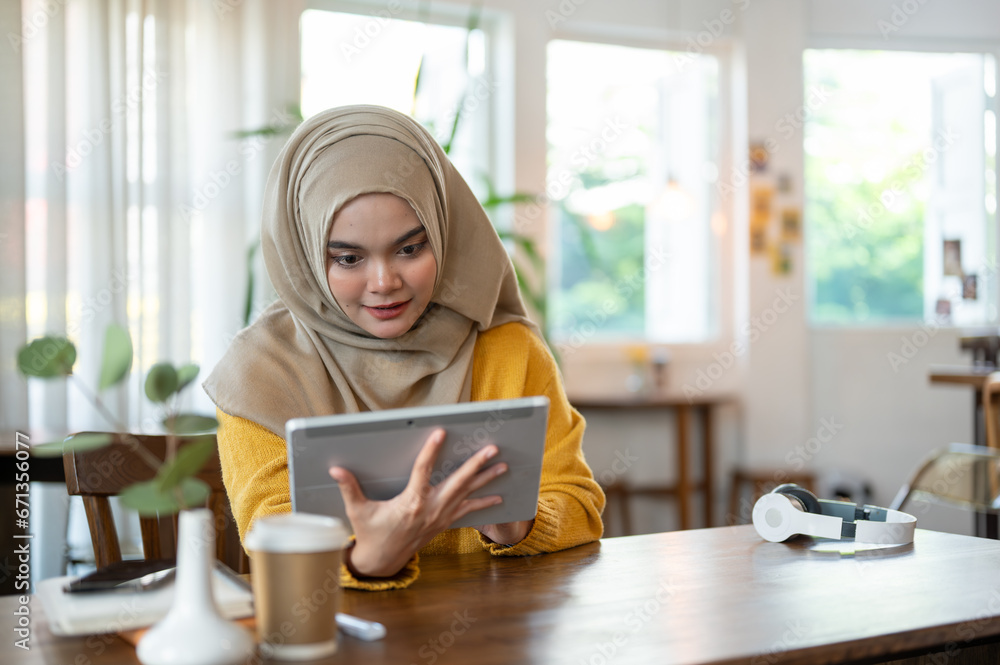 An attractive and happy Asian-Muslim woman is using a digital tablet while relaxing in a coffee shop An attractive and happy Asian-Muslim woman is using a digital tablet while relaxing in a coffee shop