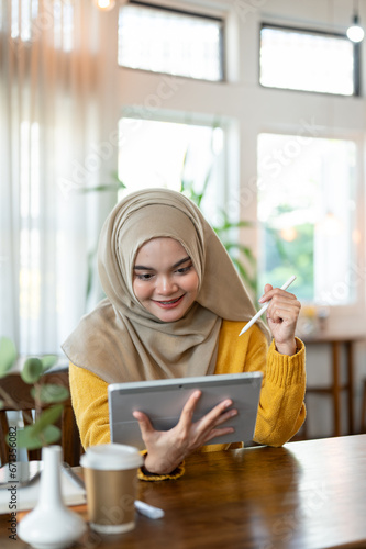 A happy Asian-Muslim woman wearing a hijab is using a tablet while relaxing in a beautiful cafe.