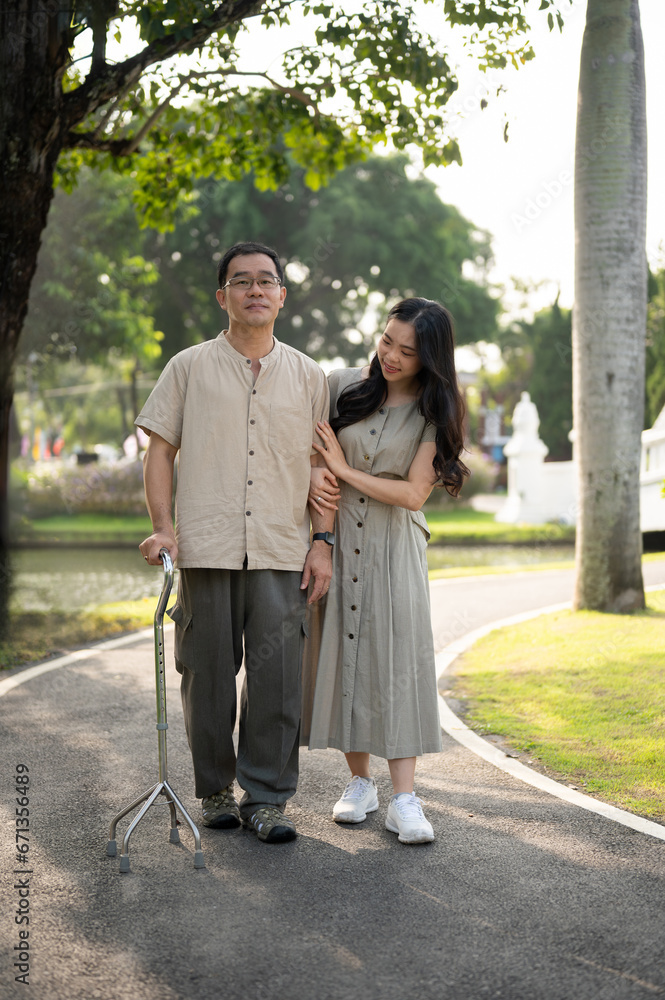 Fototapeta premium A caring young Asian daughter is walking in a green park with her senior dad in the morning.
