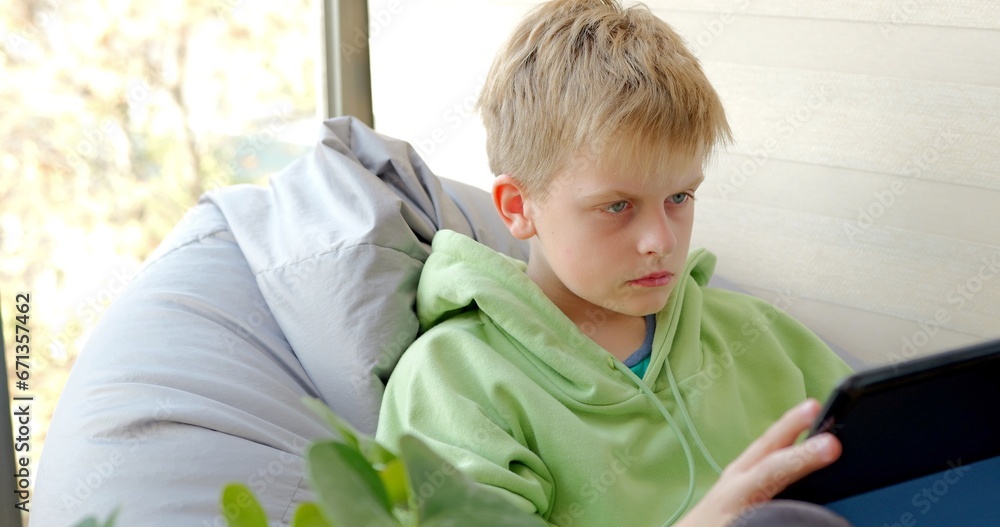 Serious boy using tablet sitting on soft chair, playing online game on tablet Serious boy using tablet sitting on soft chair, playing online game on tablet