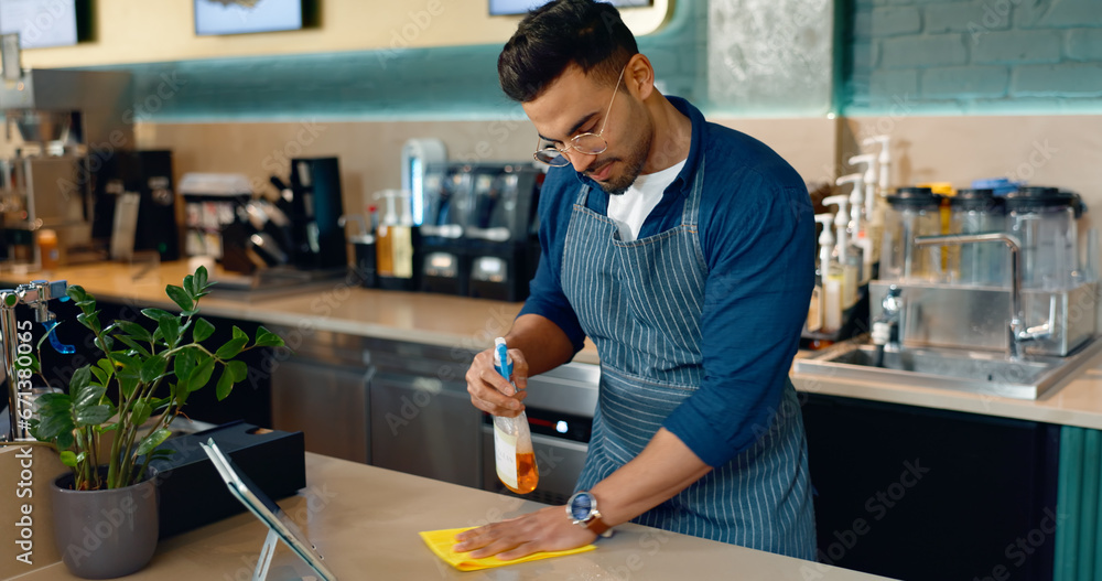 Waiter, man and cleaning table in cafe for dust, bacteria and dirt with cloth, spray or detergent. Barista, person or wipe wooden furniture in coffee shop or restaurant with chemical liquid for shine - obrazy, fototapety, plakaty 