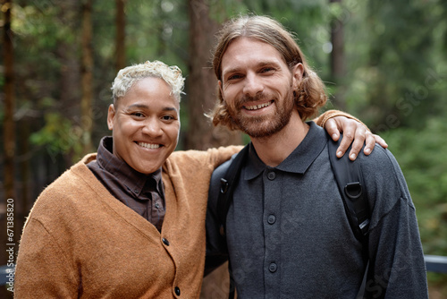 Portrait of multiethnic happy couple embracing and smiling at camera while walking in the forest