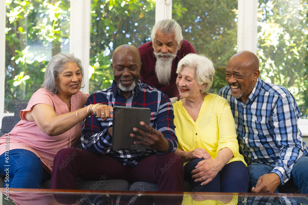 Happy diverse group of senior friends sitting on sofa, using tablet and smiling in sunny living room Happy diverse group of senior friends sitting on sofa, using tablet and smiling in sunny living room