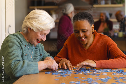 Happy diverse senior female friends playing with jigsaw puzzles in sunny dining room at home