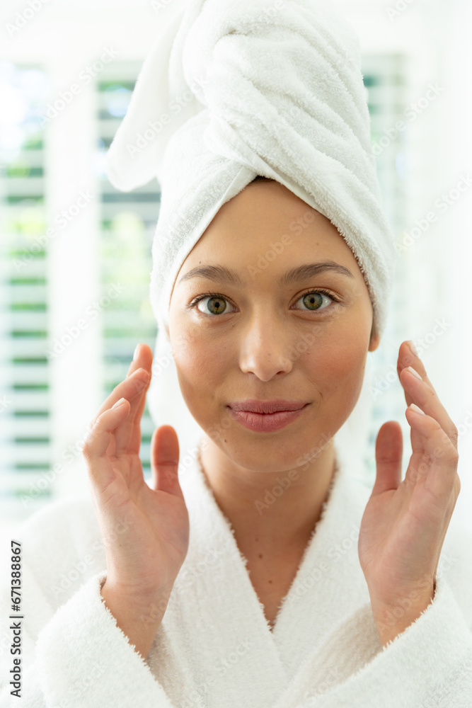 Happy caucasian woman in bathrobe looking in mirror, touching face and smiling in sunny bathroom Happy caucasian woman in bathrobe looking in mirror, touching face and smiling in sunny bathroom
