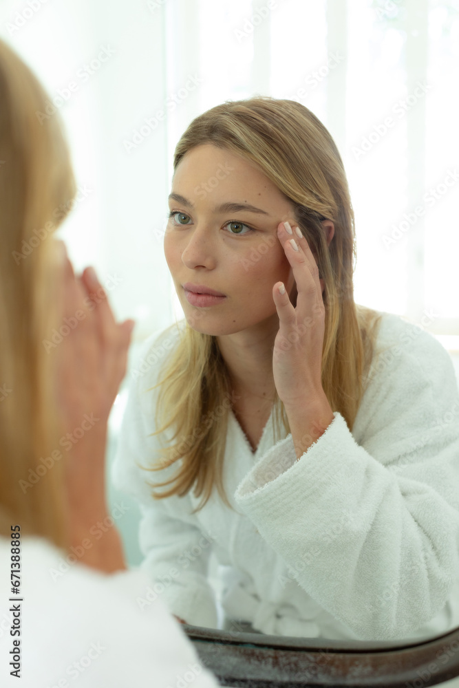 Happy caucasian woman in bathrobe looking in mirror and touching face in sunny bathroom Happy caucasian woman in bathrobe looking in mirror and touching face in sunny bathroom